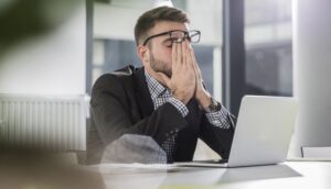 A man looking stressed while sitting at his desk, covering his face with his hands.