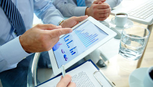 A business professional reviewing data on a tablet with colleagues in the background.