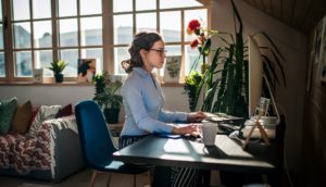 Woman working from home in a well-lit room with a plant and a comfortable workspace.
